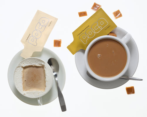 Two cups of coffee with milk foam along the edge and two poca packets on the side.