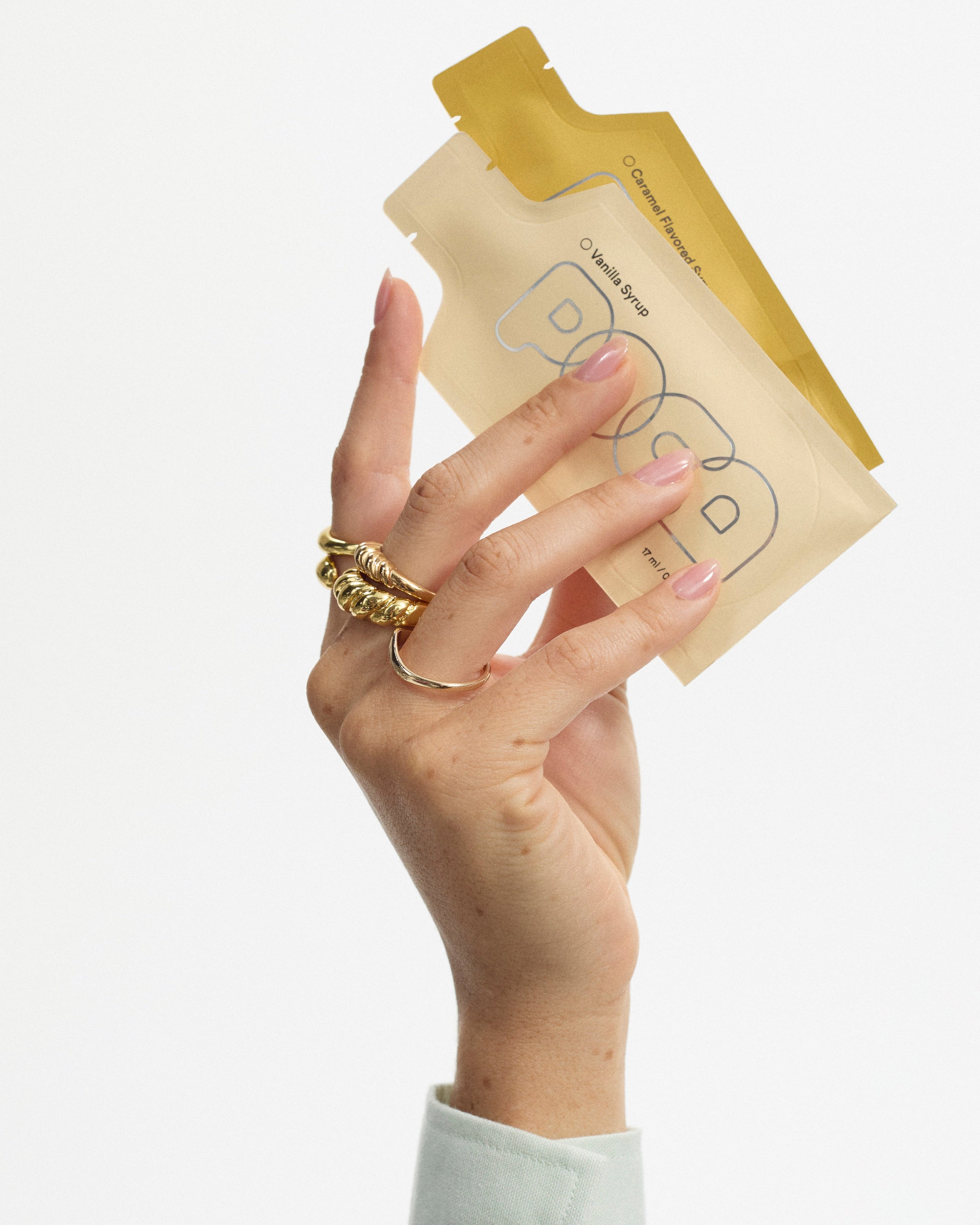 Hand with gold rings holding a vanilla and caramel POCA pouch.